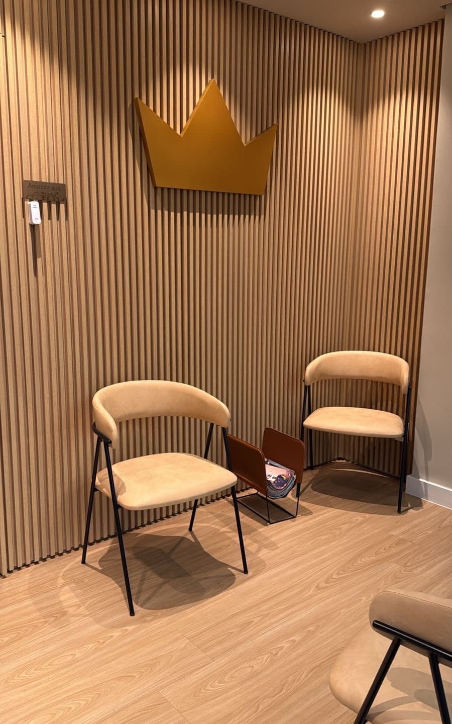 Sala de espera reservada da Ouro Savassi, com poltronas em camurça bege e a coroa dourada da marca centralizada na parede.